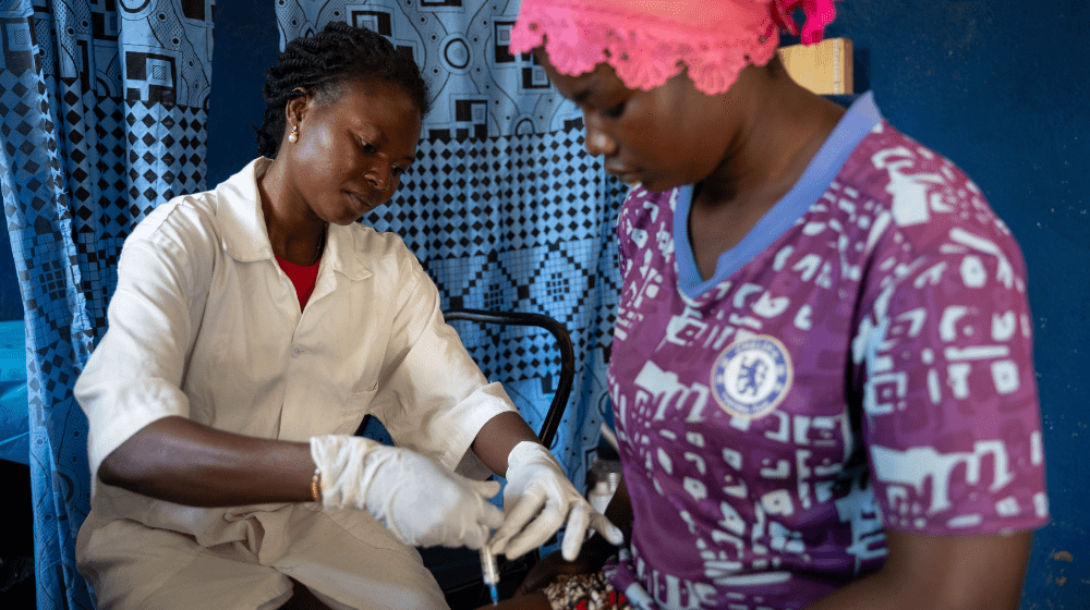 A female health worker in white uniform inserts a syringe into the leg of a female patient in a purple top and pink head wrap