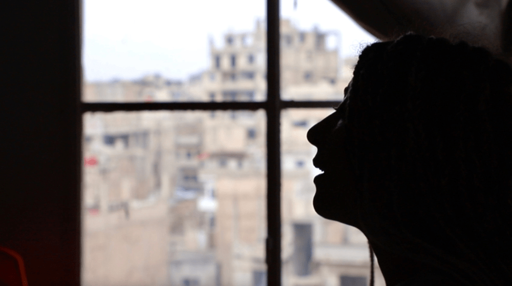  A woman stands in backlit profile beside a window, through which destroyed buildings can be seen