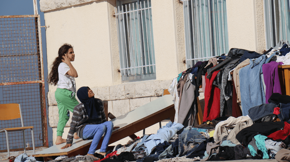  Niñas sentadas fuera de una escuela pública que sirve de refugio. Cerca hay muebles cubiertos de coloridas pilas de ropa.