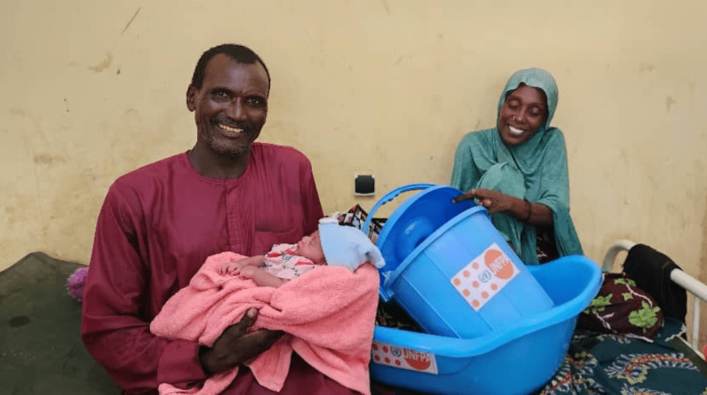 Un homme vêtu de rouge tient un nouveau-né dans ses bras. Derrière lui, la mère de l’enfant, assise sur un lit et vêtue de bleu, tient une petite baignoire bleue avec le logo de l’UNFPA.