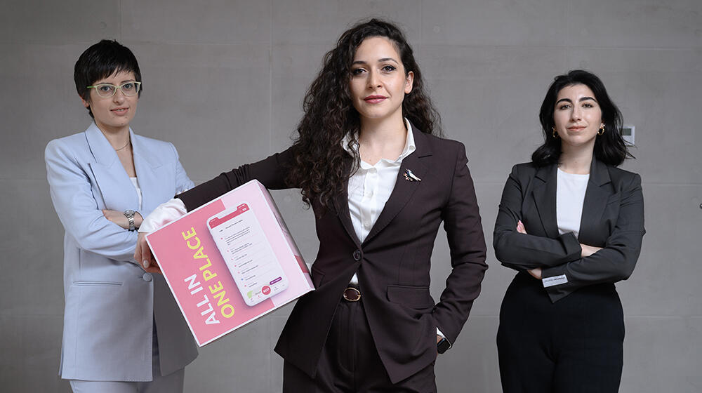 Three woman stand together against a gray wall. One of them is holding a poster.