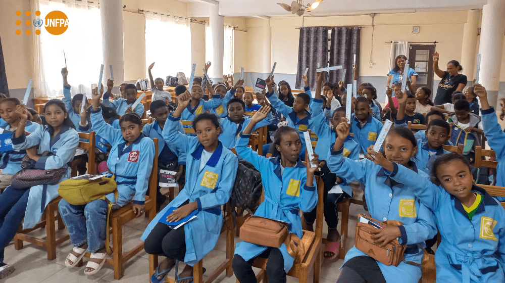 Un groupe de jeunes élèves en uniformes bleus lèvent la main, assis·e·s dans une salle de classe