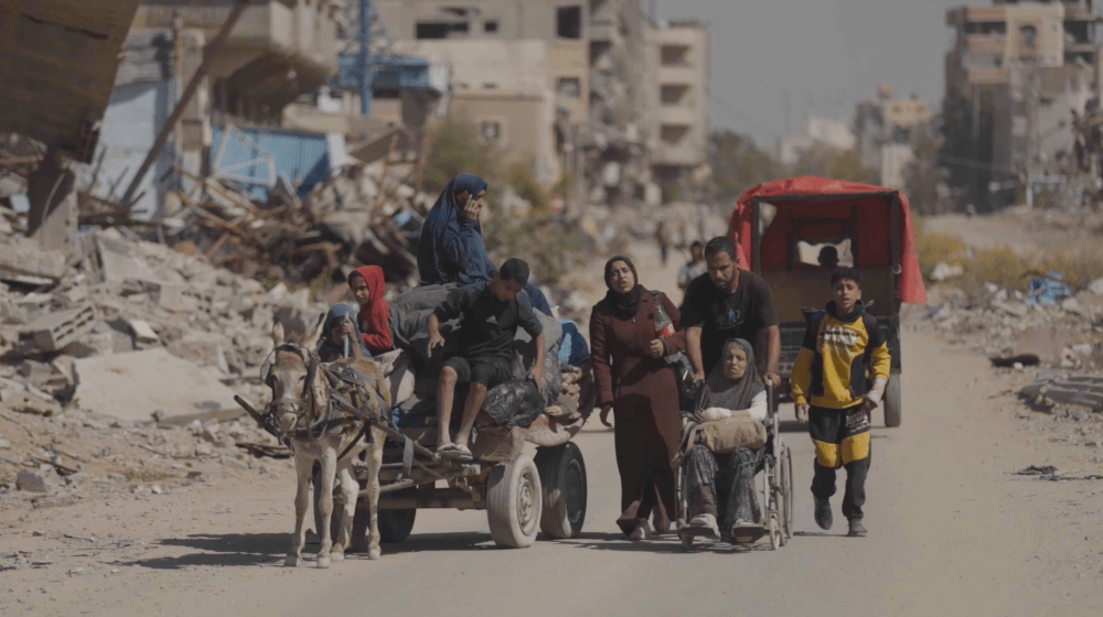 Un groupe de personnes, dont une en fauteuil roulant et plusieurs sur une carriole tirée par un âne, s’avancent sur une route poussiéreuse encadrée de bâtiments en ruines et de gravats