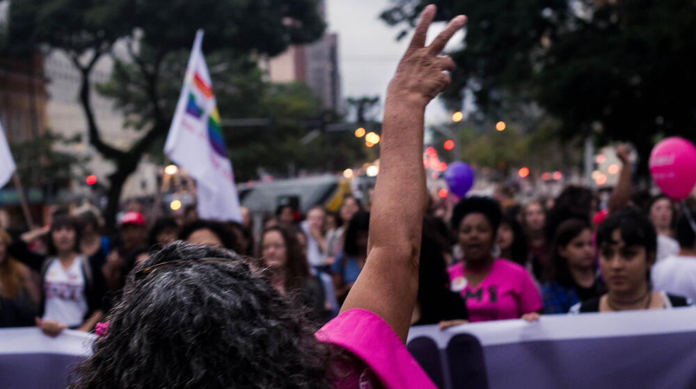 A protester faces a crowd who are demonstrating. She forms a 'peace' sign with her hand.