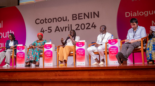 Five global youth dialogue speakers sit on a panel on a stage, one of them is holding a microphone and speaking.