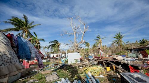 A man stands among broken buildings, trees and water.