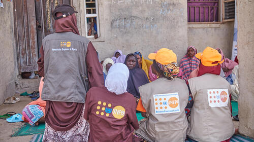 Women with the UNFPA logo embroidered on their clothes speak to a group of women sitting on blankets in a courtyard.