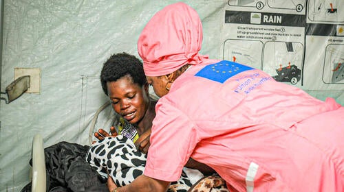  A midwife in pink uniform leans over a woman holding a baby, lying on a bed in a tent